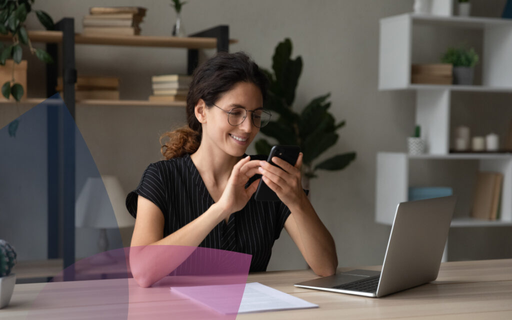 A woman scrolling her phone, sitting at a desk in front of an open laptop